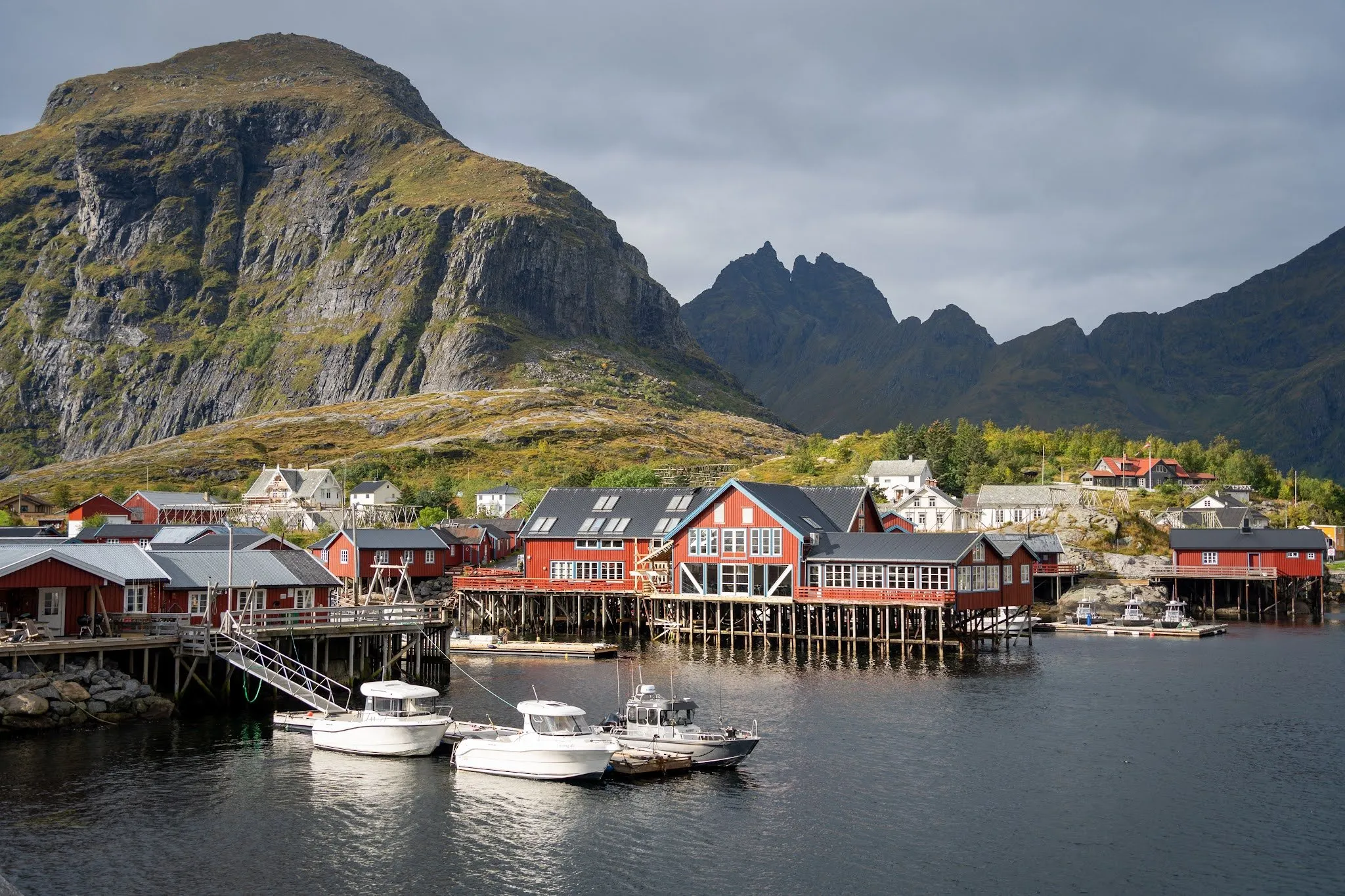 Rote Fischerhäuser auf Stelzen und weisse Boote im Hafen von Å, dahinter ein steiler Berg und ein gezackter Bergkamm