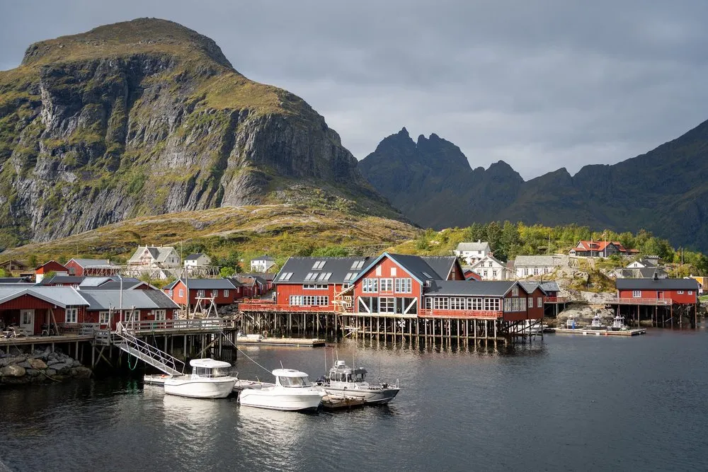 Rote Fischerhäuser auf Stelzen und weisse Boote im Hafen von Å, dahinter ein steiler Berg und ein gezackter Bergkamm