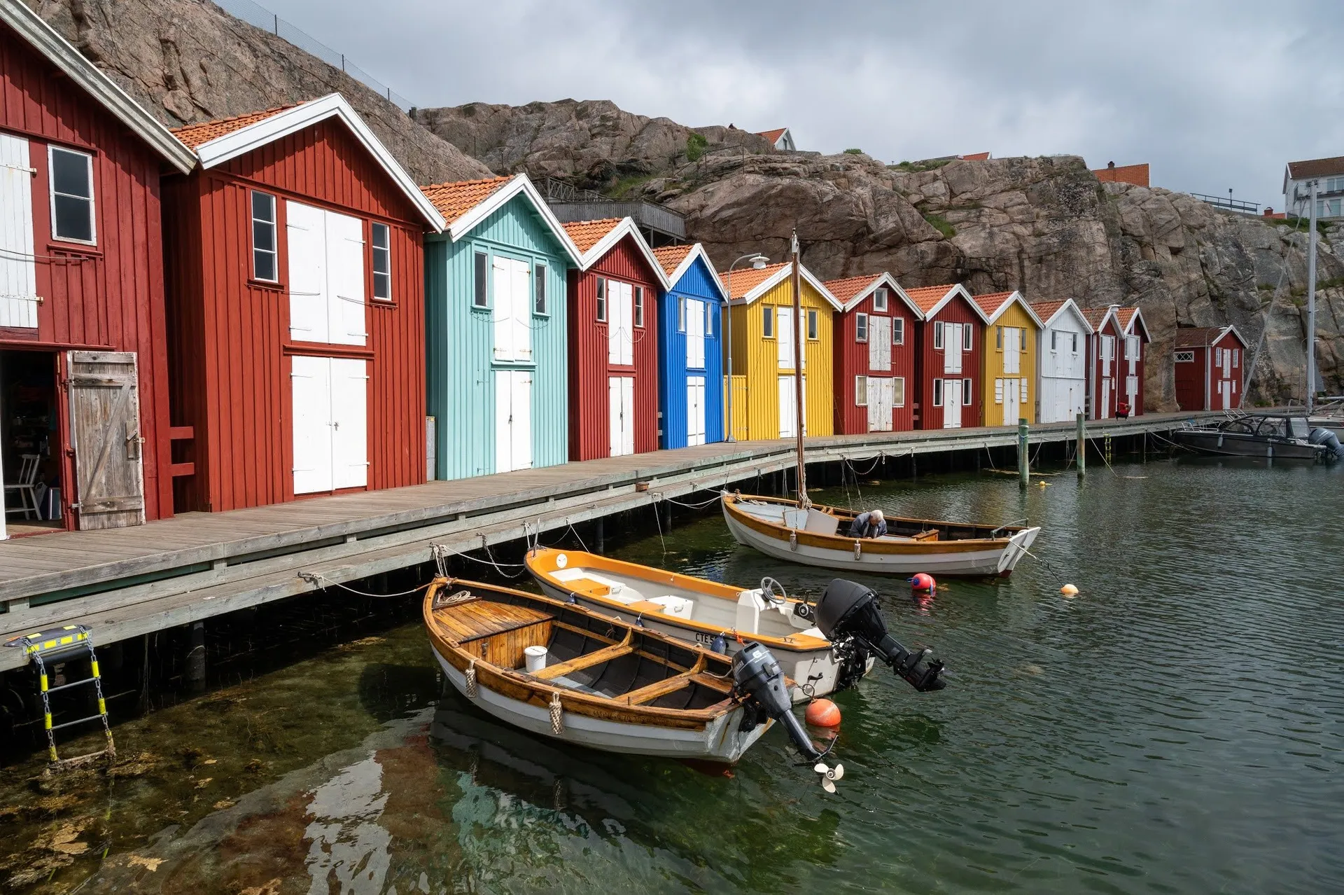 A wooden jetty with red, blue and yellow huts, three small boats in the clear water in front