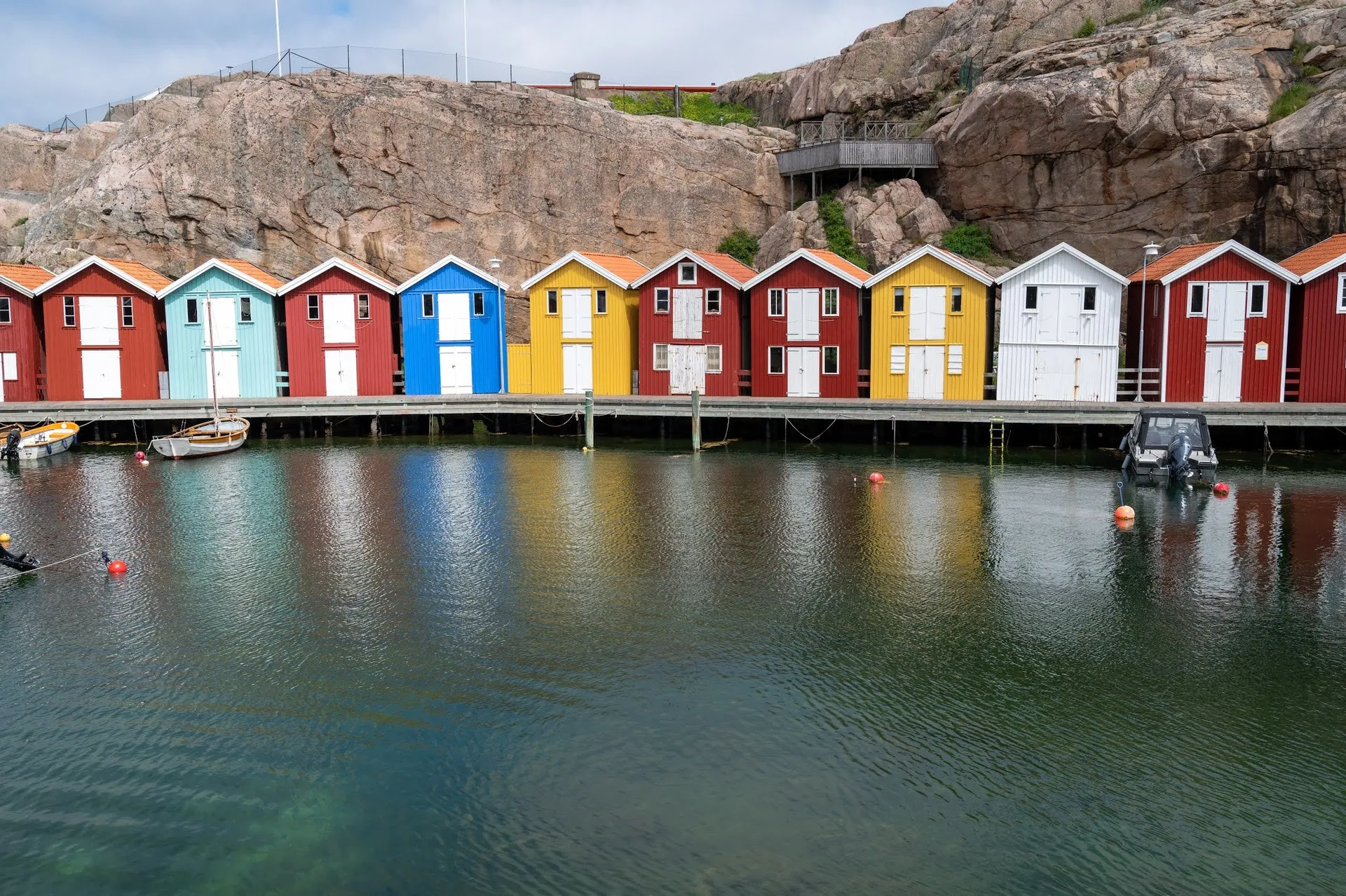 A row of red, blue, yellow and white huts on the water in front of a rock face, reflected in the water