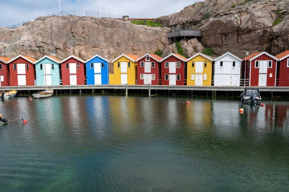 Eine Reihe roter, blauer, gelber und weisser Hütten am Wasser vor einer Felswand, gespiegelt im Wasser
