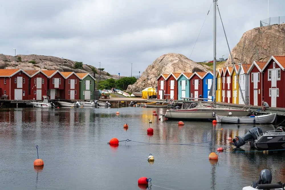 Rote und einzelne bunte Hütten hinter einem Hafenbecken mit orangen Bojen, einem Segelmast und einem Felsen