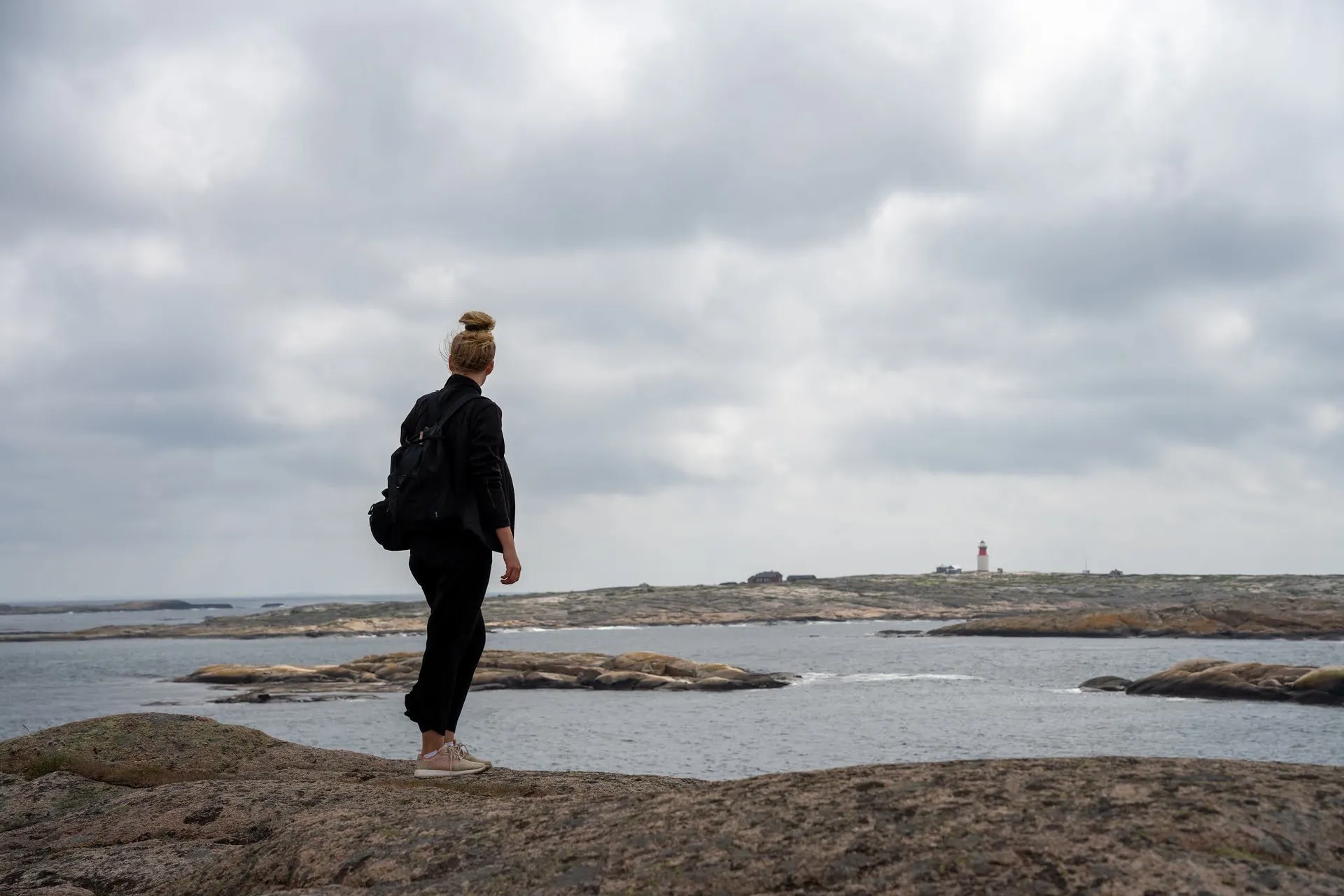 A woman with a backpack stands on the rock and looks out to sea, a small lighthouse on the horizon