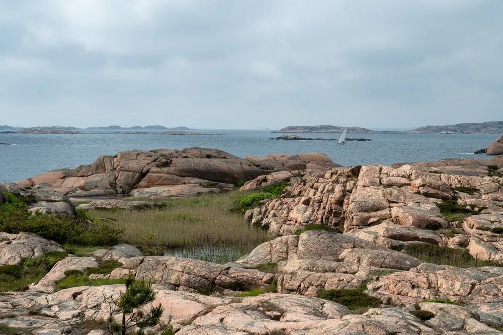 Rötliche Granitfelsen mit Grasflächen, dahinter das Meer und ein Segelboot