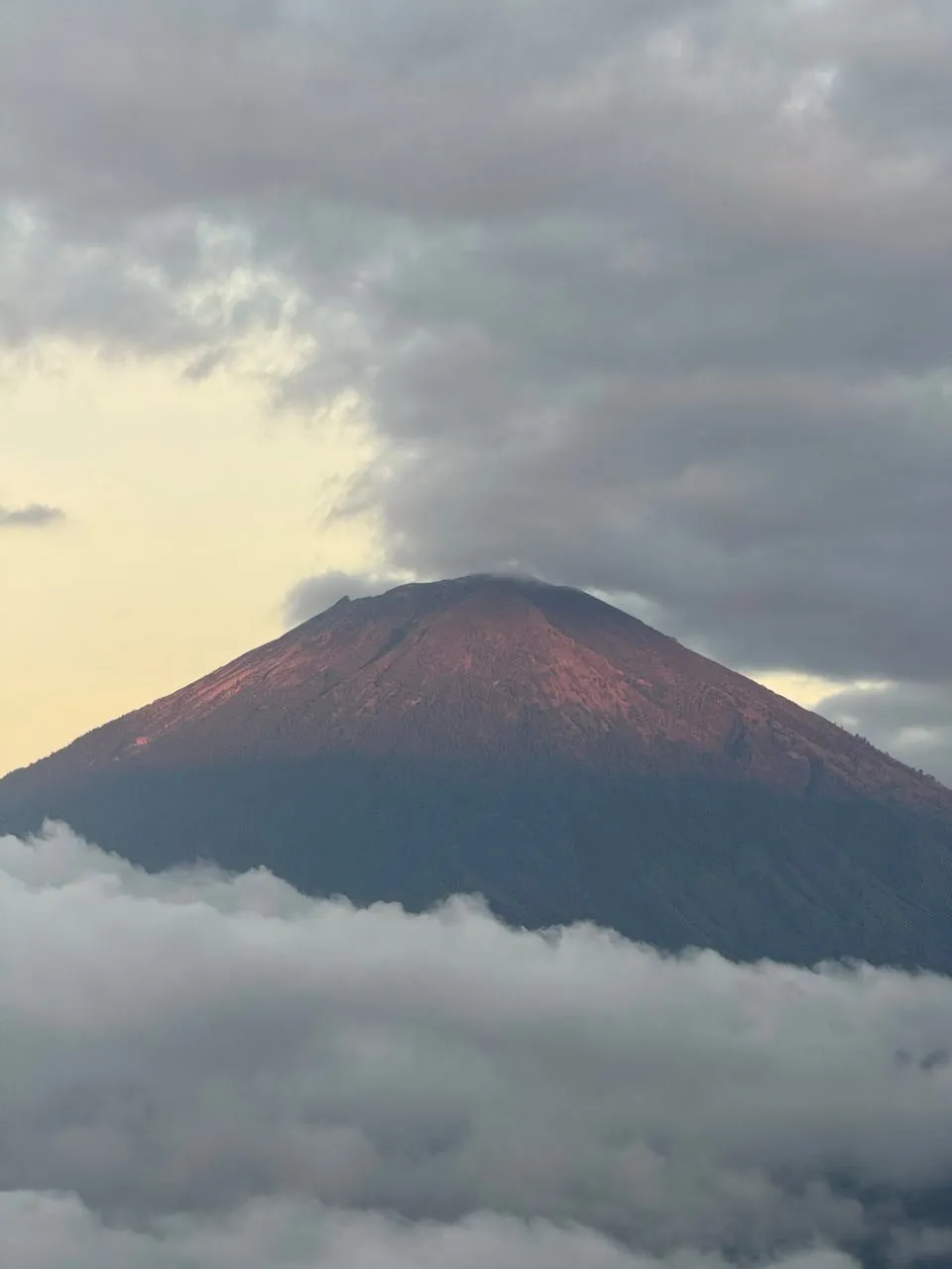 Der rötlich beleuchtete Kegel des Mount Agung über einem Wolkenband, um den Gipfel hängen graue Wolken
