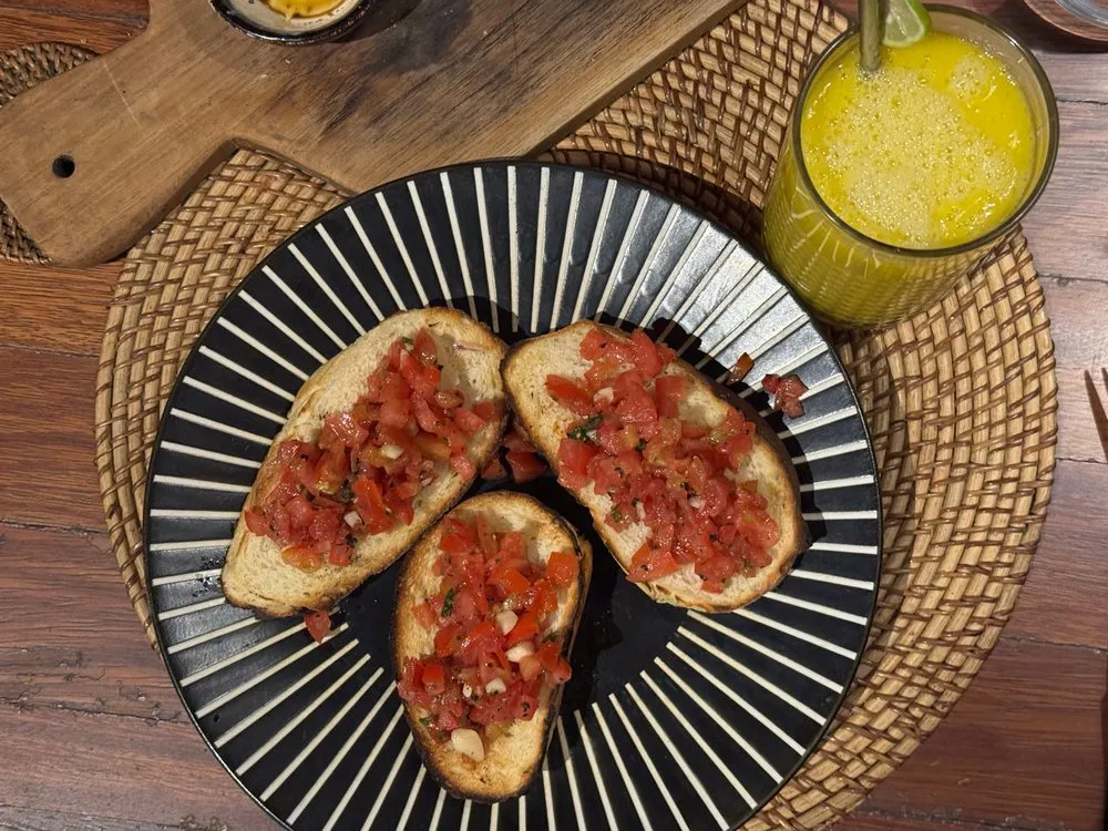 Bruschetta mit gehackten Tomaten auf einem schwarz-weiss gestreiften Teller, daneben ein gelbes Getränk mit Limettenscheibe