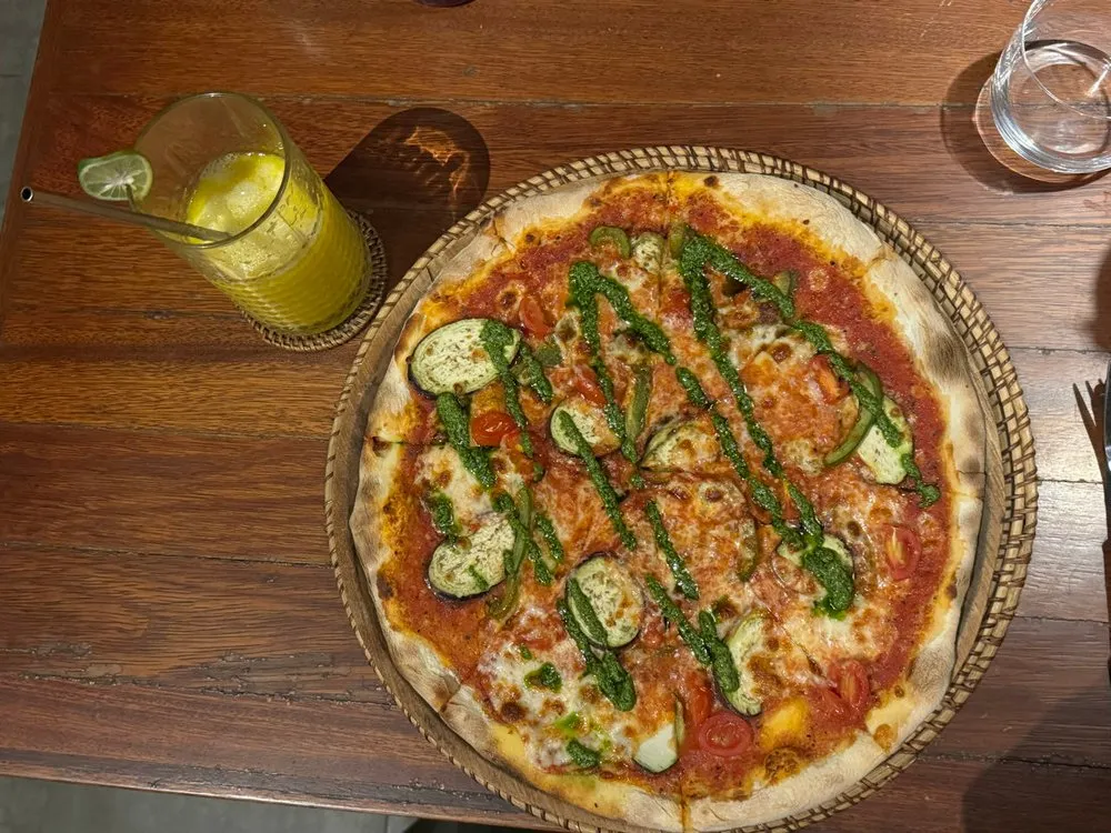 Pizza mit Tomatensauce, Käse, Gemüsescheiben und grünem Pesto auf einem geflochtenen Tablett, daneben ein gelbes Getränk