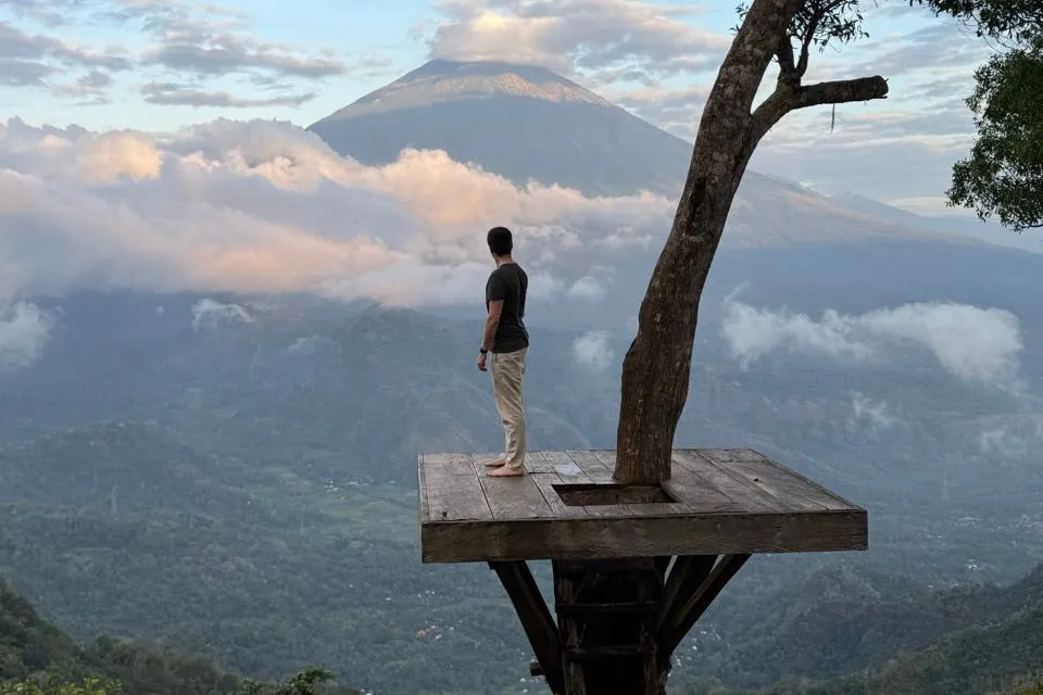 Nicolas steht barfuss auf einer Holzplattform um einen Baum und blickt über ein Tal mit rosa beleuchteten Wolken auf den Vulkan Mount Agung
