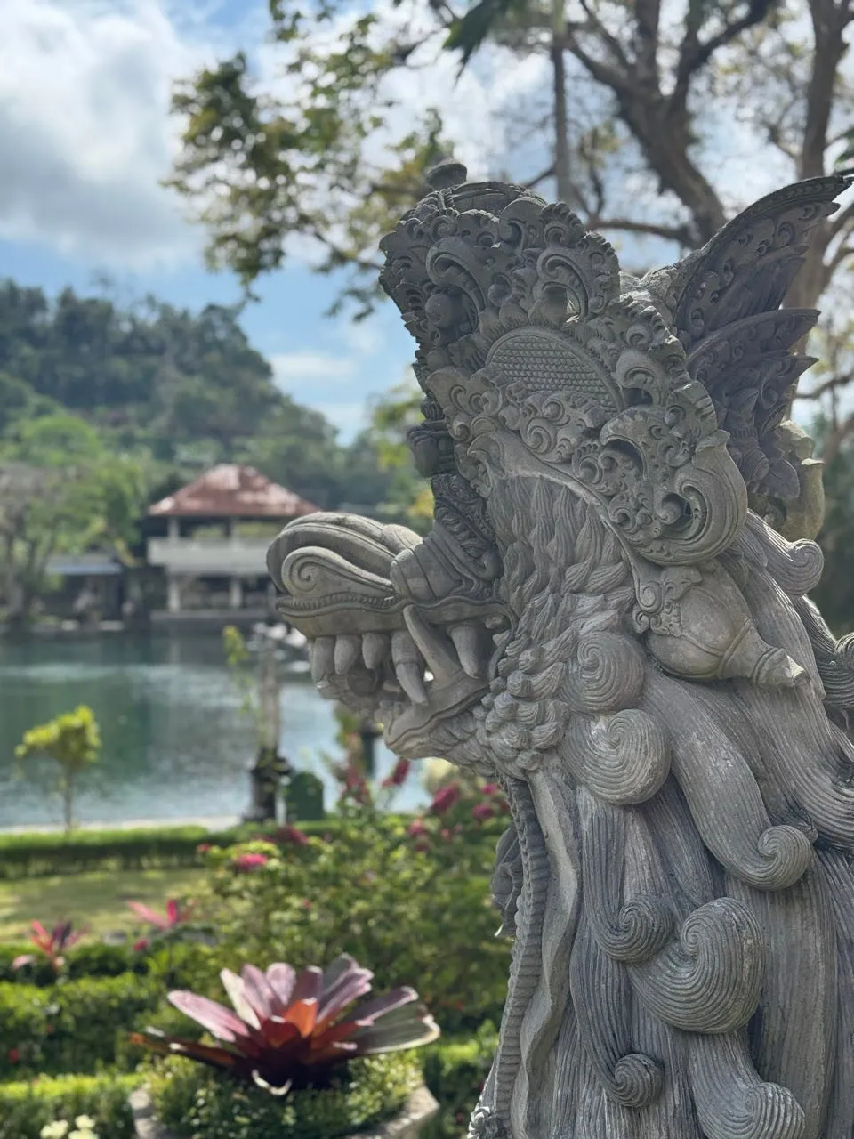 Der Kopf einer Steinfigur mit Fangzähnen, dahinter ein Teich und ein Pavillon
