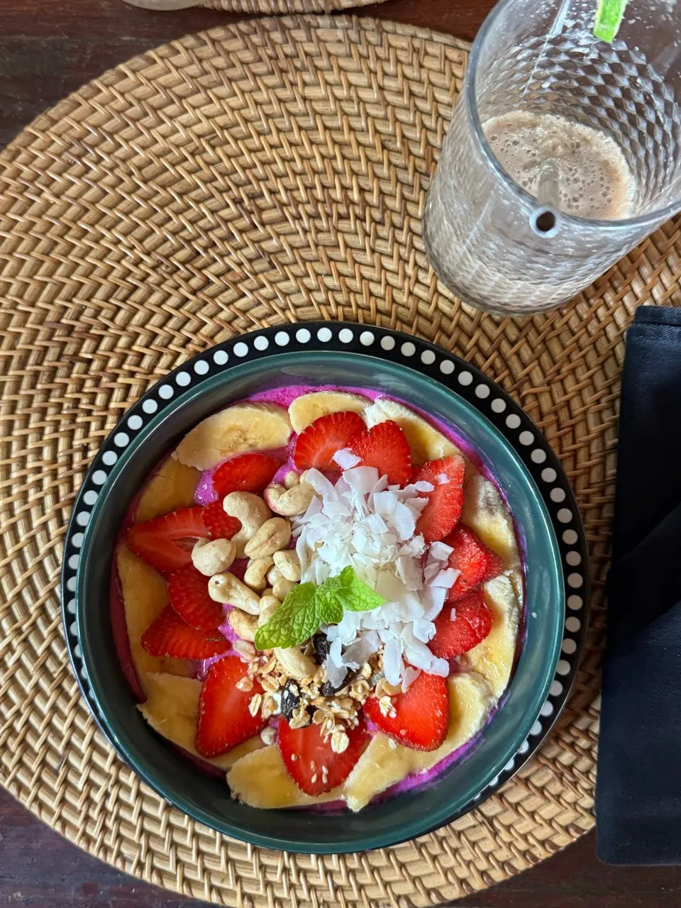 Frühstücksbowl mit Erdbeeren, Bananenscheiben, Cashewkernen und Kokosflocken auf einem Bastset, daneben ein Glas mit hellbraunem Getränk