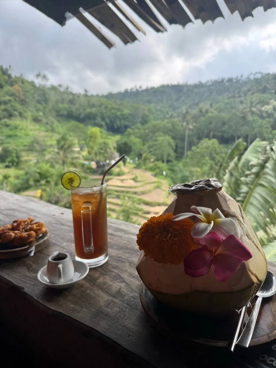 Auf dem Holztisch stehen frittierte Banane, ein Glas mit Limettenscheibe und eine mit Blüten geschmückte Kokosnuss, dahinter die Terrassen