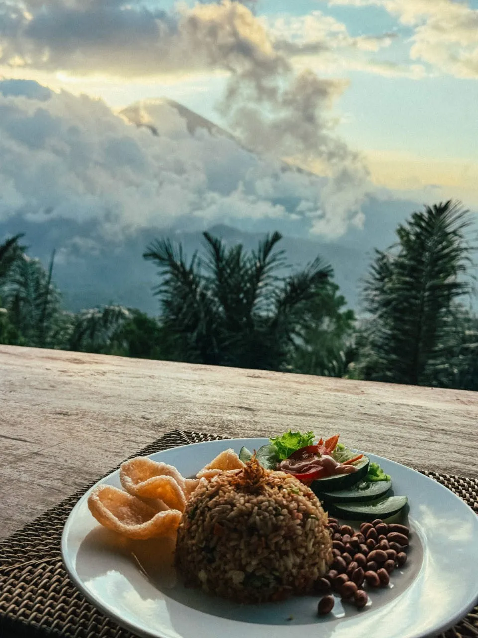 Ein Teller mit gebratenem Reis, Krupuk und Gurkenscheiben, dahinter Wolken und ein Berggipfel im Abendlicht