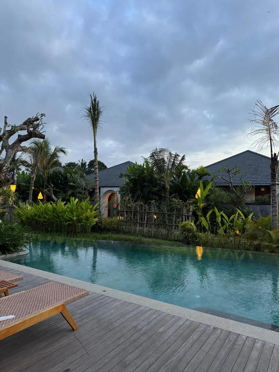 Der Pool in der Dämmerung mit Holzdeck, Liegen, Palmen und Häusern mit dunklen Dächern