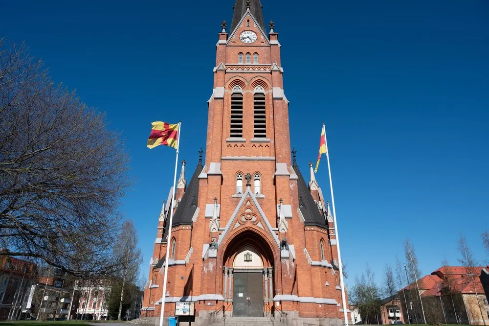 Die Kathedrale von Luleå aus rotem Backstein mit hohem Turm und zwei wehenden Fahnen vor tiefblauem Himmel