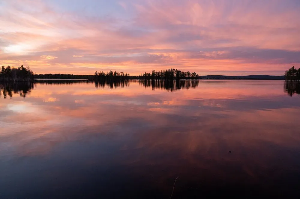 Rosa und orange Wolken spiegeln sich im spiegelglatten See, am Ufer stehen Baumsilhouetten