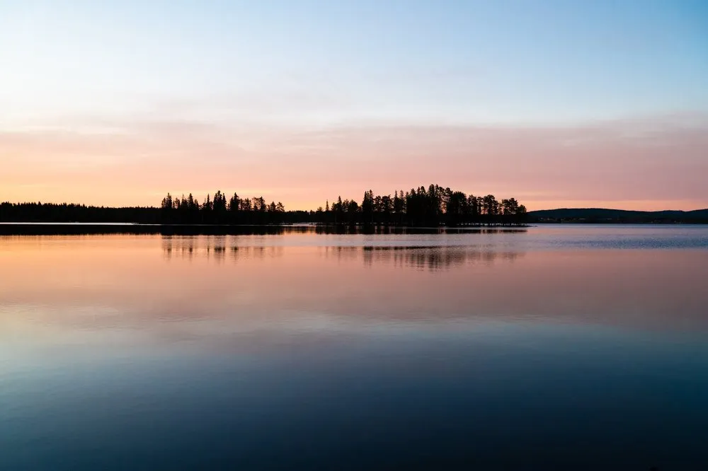 Blasser Abendhimmel in Rosa und Blau, eine bewaldete Landzunge spiegelt sich im ruhigen See
