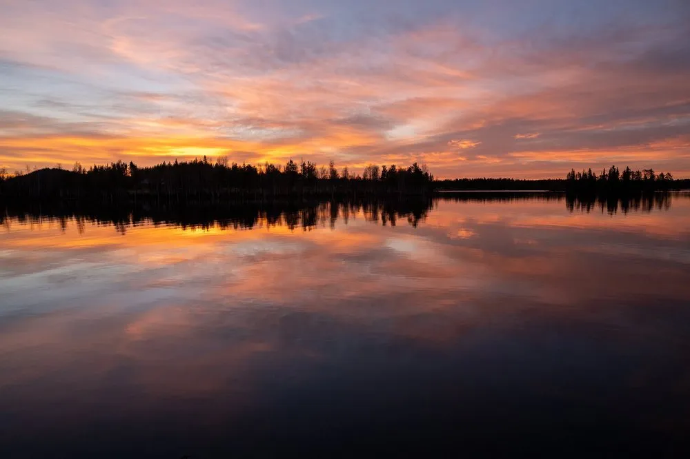Orange und violette Wolken über dem See, die Baumreihe am Ufer spiegelt sich im dunklen Wasser