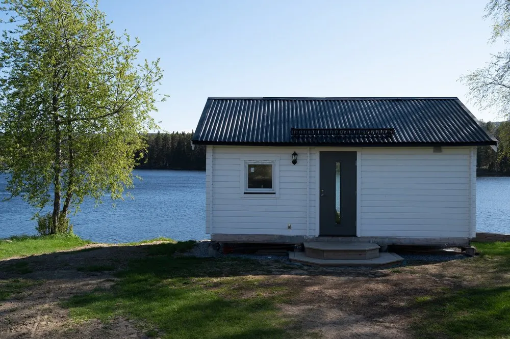 Ein kleines weisses Häuschen mit dunklem Dach steht auf einer Wiese direkt am blauen See