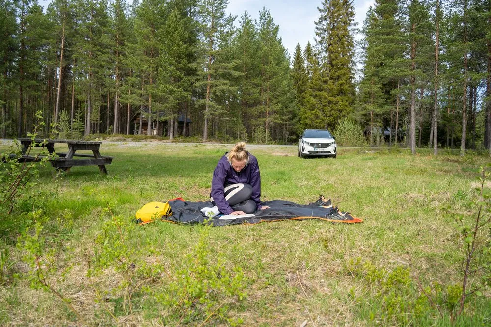 Eine Frau sitzt auf einer Picknickdecke im Gras und liest, dahinter ein Picknicktisch, Wald und ein weisses Auto