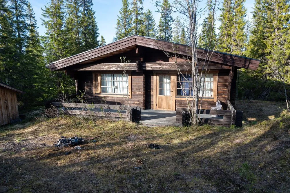 Das dunkelbraune Blockhaus des Airbnb im Wald mit Veranda, links ein kleiner Holzschuppen