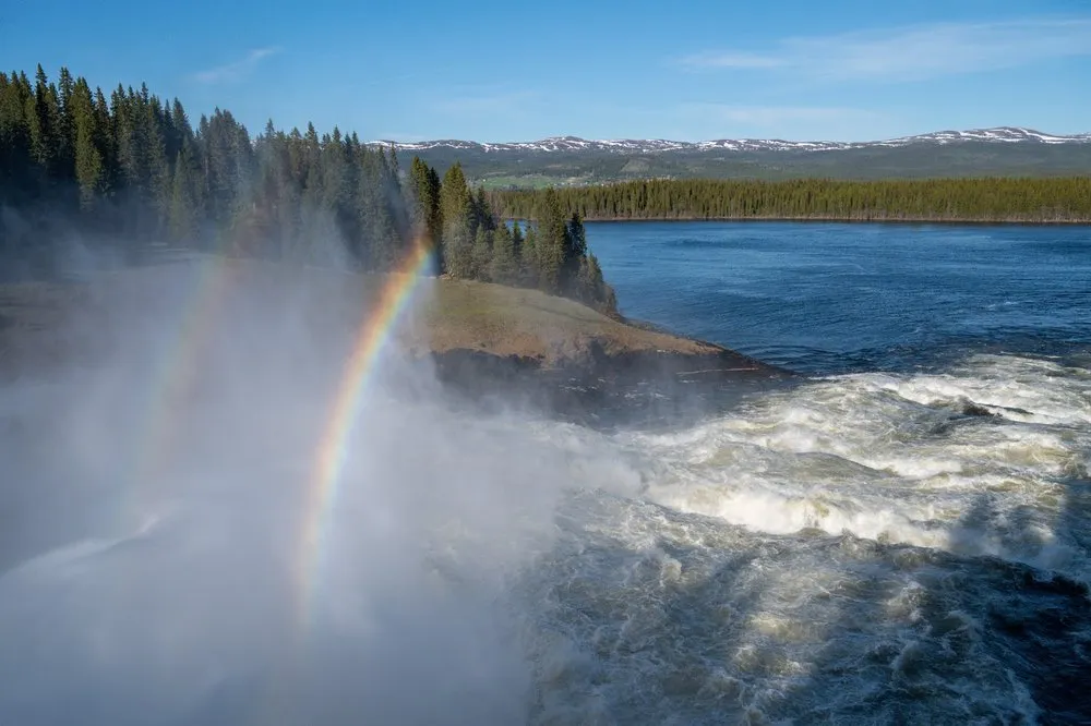 Doppelter Regenbogen in der Gischt des Tännforsen, dahinter Wald, ein See und Berge mit Schneeflecken