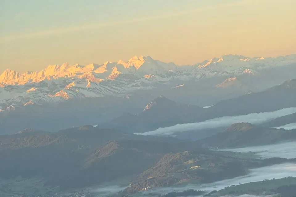 Orange beleuchtete Schneegipfel der Alpen über dunklen Hügeln und Nebelbändern im Morgenlicht