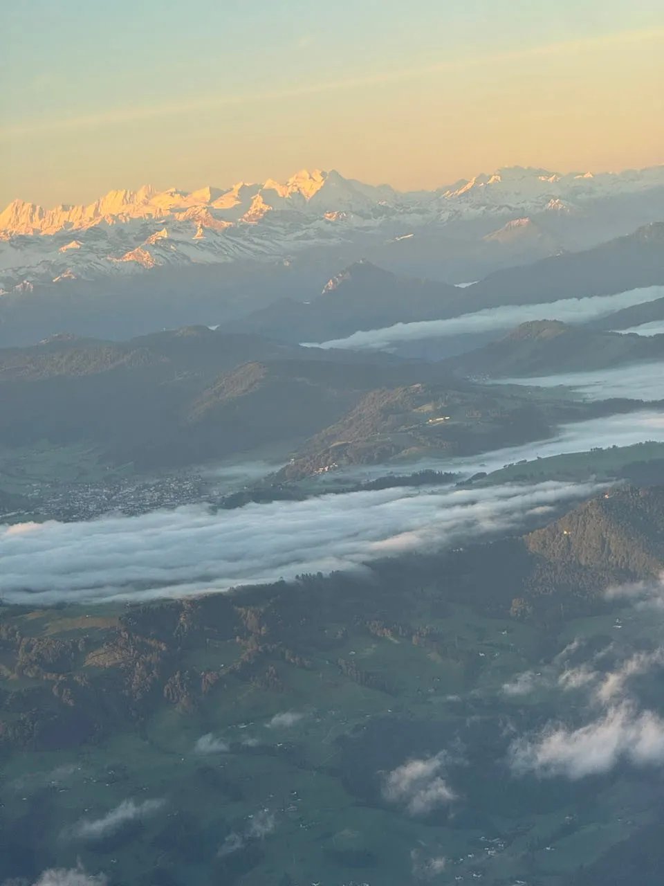 Blick aus dem Flugzeug auf verschneite Alpengipfel, davor dunkle Hügel, Nebelbänke und grüne Täler mit einzelnen Siedlungen