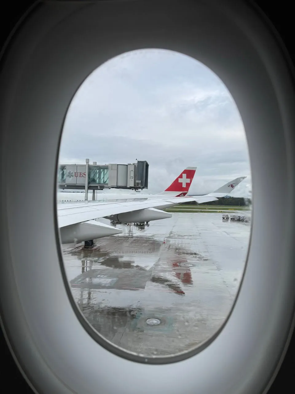 Blick aus dem Flugzeugfenster auf den nassen Asphalt, eine Fluggastbrücke und ein rotes Heck mit weissem Kreuz