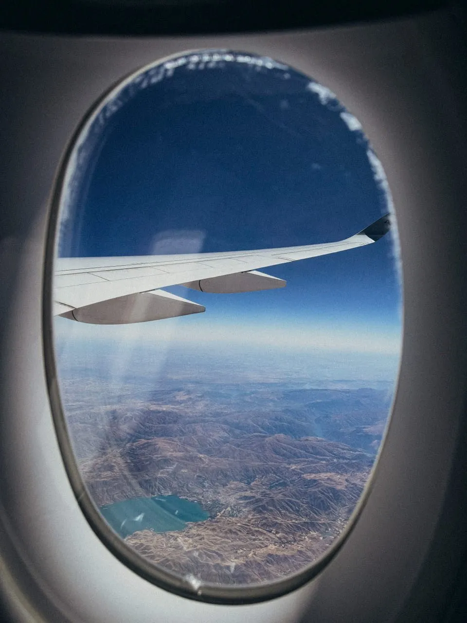 Flugzeugflügel über braunen Bergen, am unteren Bildrand ein türkisfarbener See