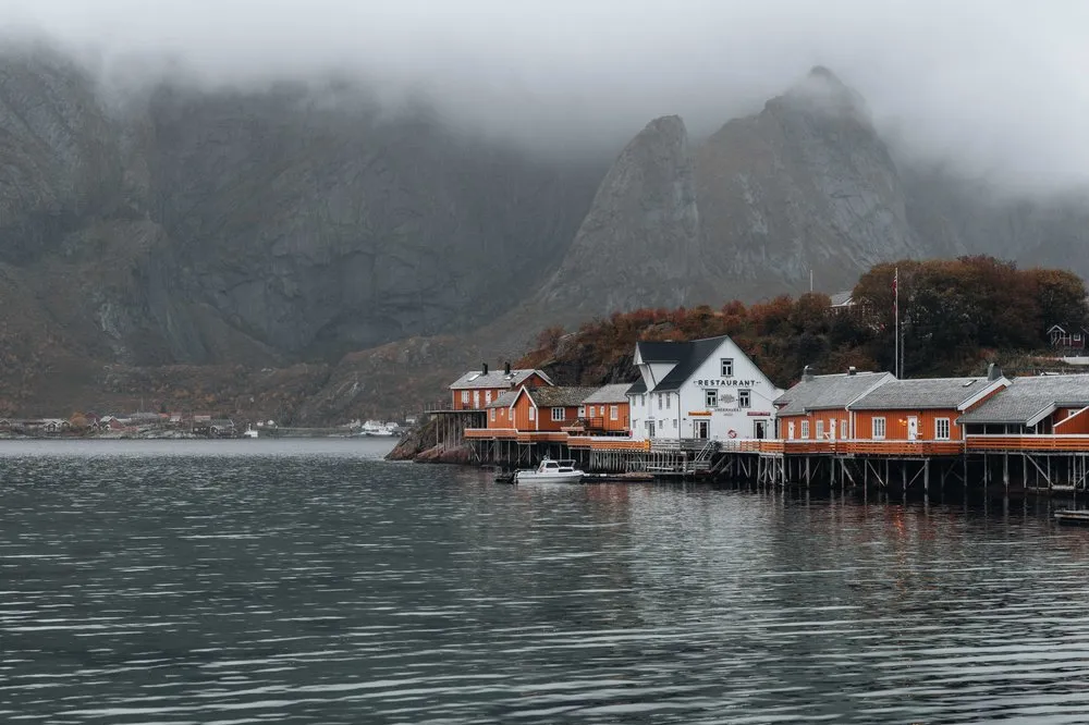 Die ockergelben Rorbuer von Sakrisøy auf Pfählen über dem Wasser, dahinter die Berge in tiefen Wolken