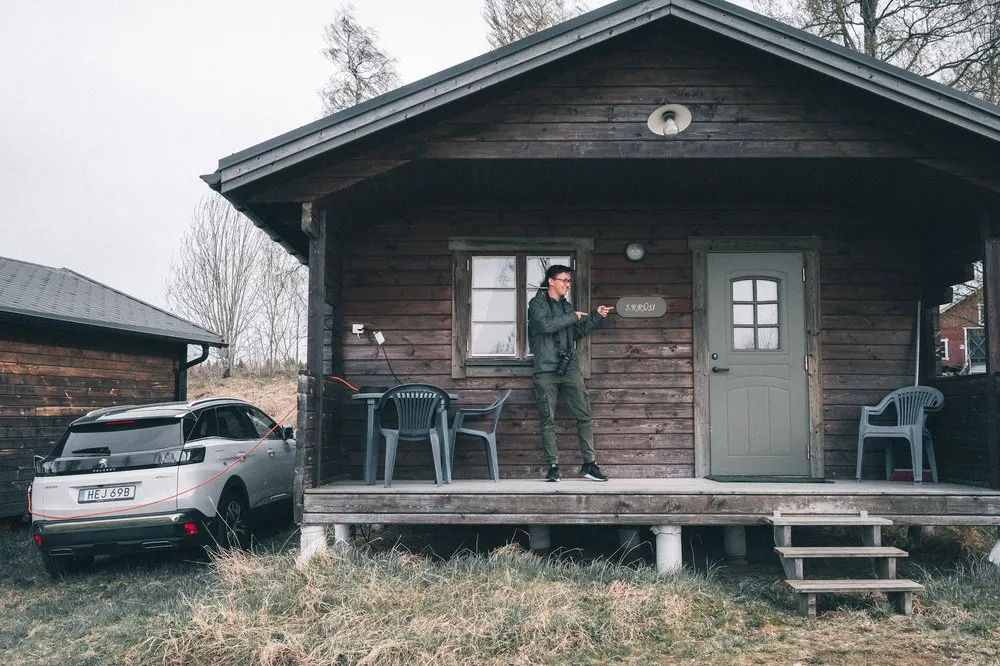 Das Häuschen aus dunklem Holz mit Veranda, ein Mann zeigt auf das Namensschild neben der Tür, links steht ein weisses Auto
