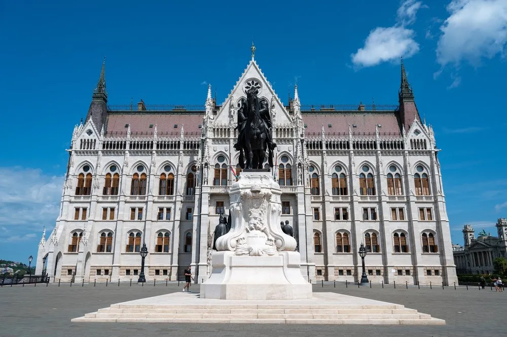 Frontansicht des Parlamentsgebäudes mit Reiterstatue auf einem hellen Sockel, davor ein weiter Platz unter blauem Himmel
