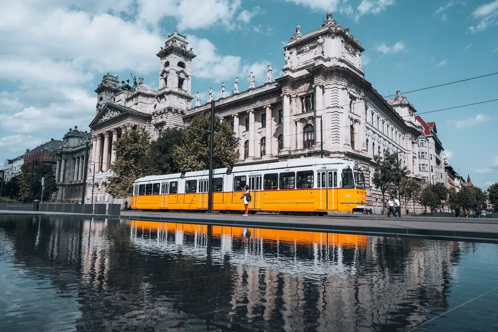 Eine gelbe Strassenbahn fährt an einem hellen Gebäude mit Turm vorbei, das sich im Wasser davor spiegelt