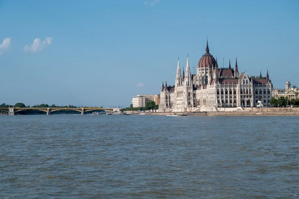 Blick über die Donau auf ein weisses Gebäude mit roter Kuppel und Spitztürmen am Ufer, links eine Brücke