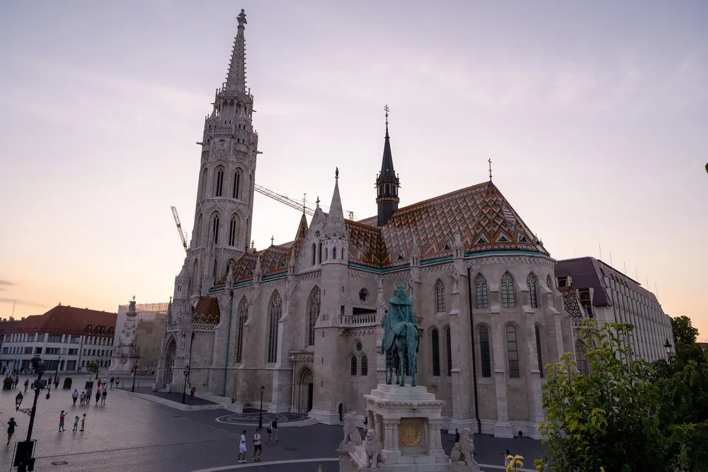 Die Kirche mit hohem Turm und buntem Ziegeldach im Abendlicht, davor eine Reiterstatue auf dem Platz