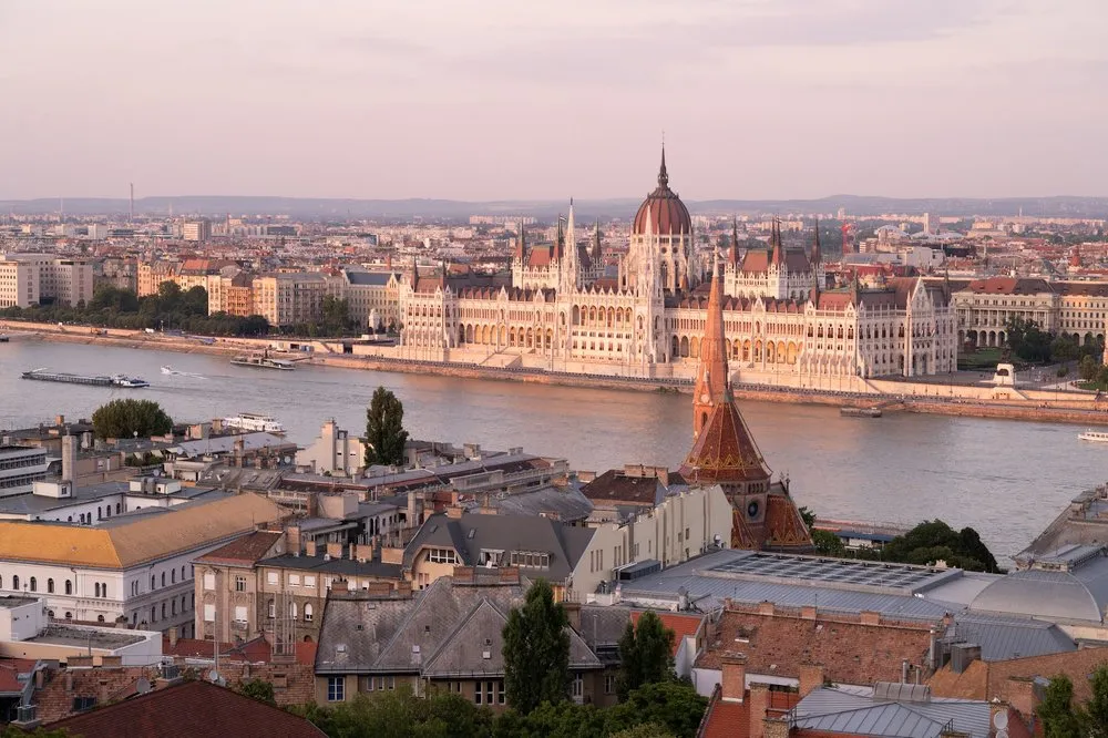 Blick über Dächer und die Donau auf ein langes Gebäude mit Kuppel am Ufer der Pest Seite im warmen Abendlicht
