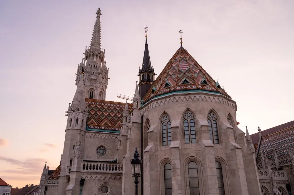 Das gemusterte Ziegeldach und der Turm der Kirche von unten vor rosa Himmel, davor eine Strassenlaterne