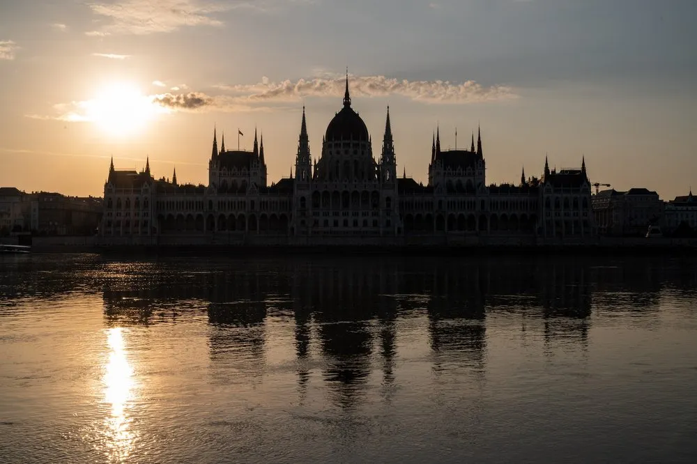 Das Parlament als Silhouette im Morgenrot, links die aufgehende Sonne und ihre Spiegelung in der Donau