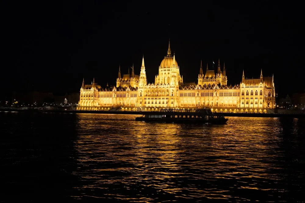 Das beleuchtete Parlamentsgebäude von Budapest spiegelt sich bei Nacht in der Donau, davor ein Ausflugsschiff