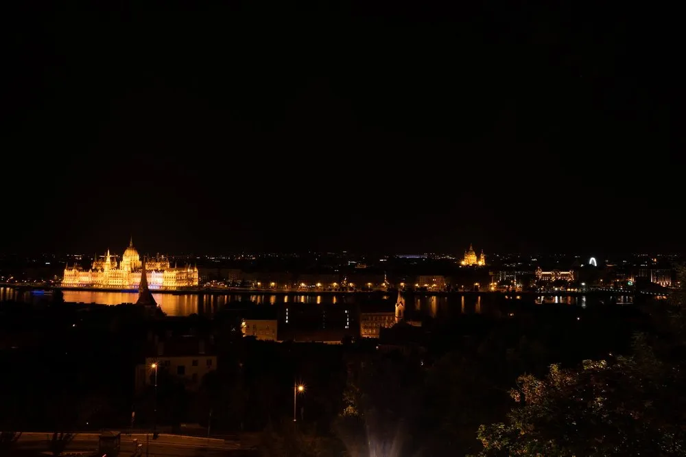 Nachtaufnahme von oben über den dunklen Fluss, am anderen Ufer leuchtet ein Gebäude mit Kuppel neben weiteren Lichtern