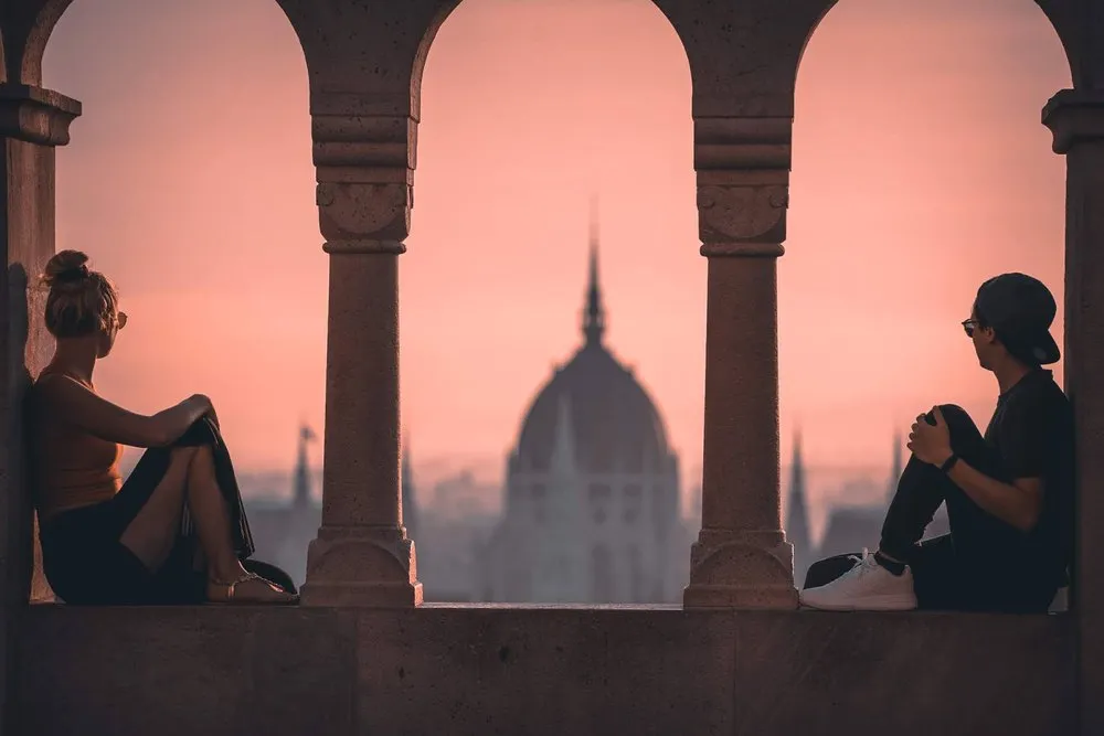 Zwei Personen sitzen auf der Brüstung der Fisherman's Bastion, zwischen den Säulen die Kuppel des Parlaments im rosa Morgenlicht
