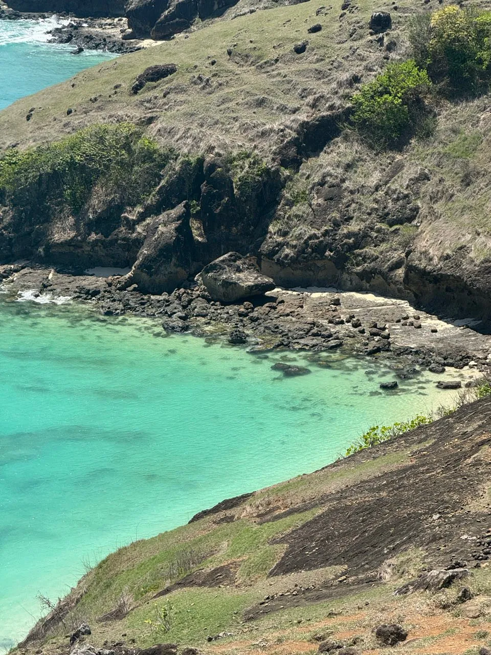 Kleine Felsenbucht mit türkisfarbenem Wasser, dunklen Felsen, einem schmalen Sandstreifen und grasbewachsenem Hang
