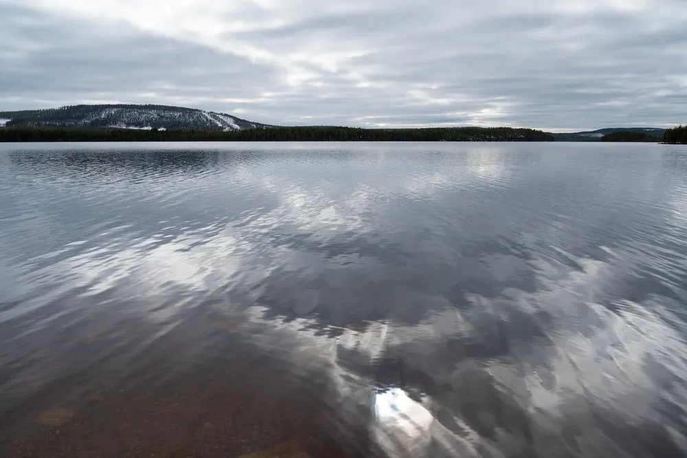 Wolken spiegeln sich auf dem ruhigen, offenen Wasser eines Sees, am Ufer ein bewaldeter Hügel mit Schneeresten