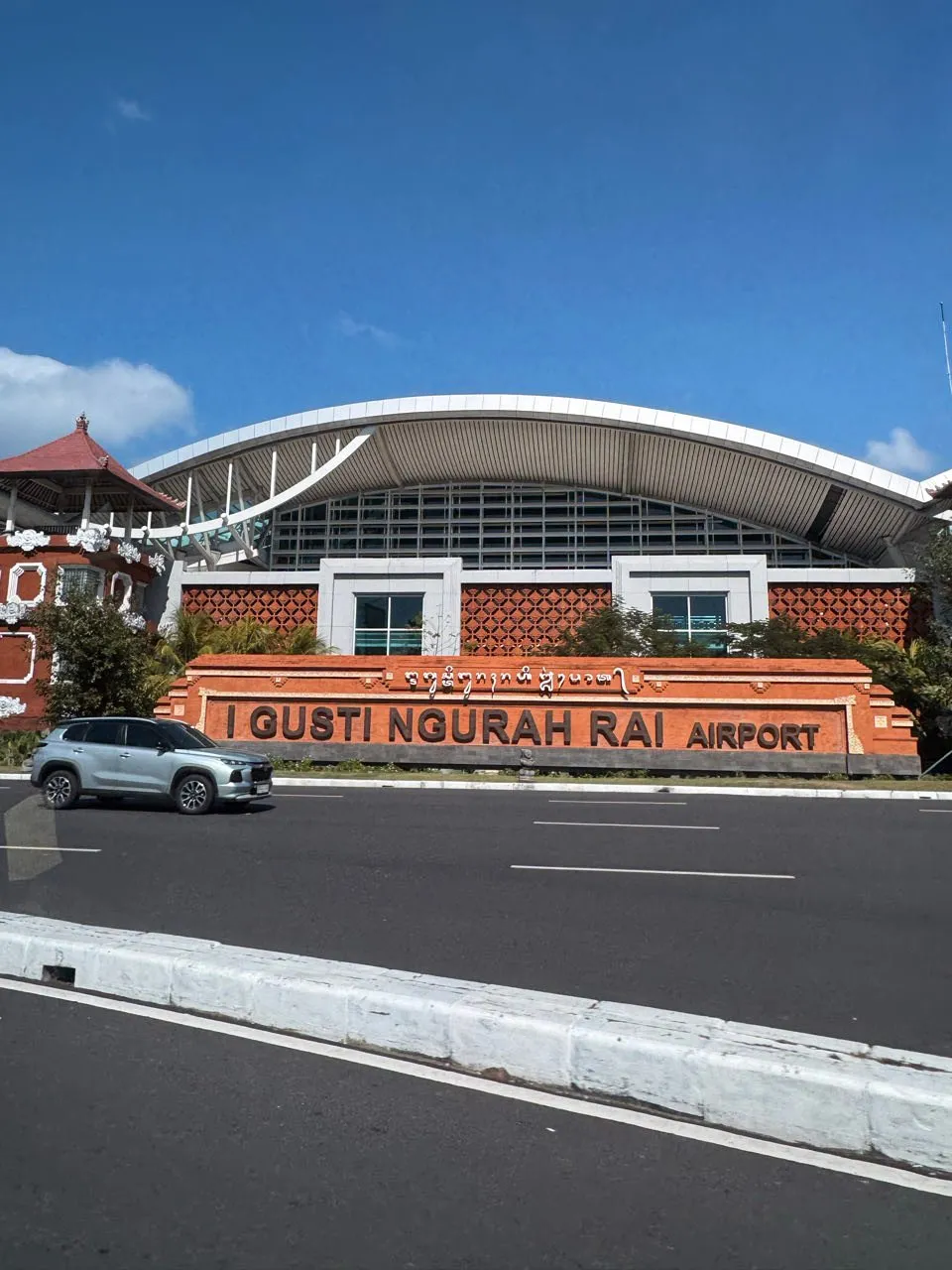 Der Eingang des Flughafens I Gusti Ngurah Rai mit rotbrauner Fassade und geschwungenem weissem Dach unter blauem Himmel
