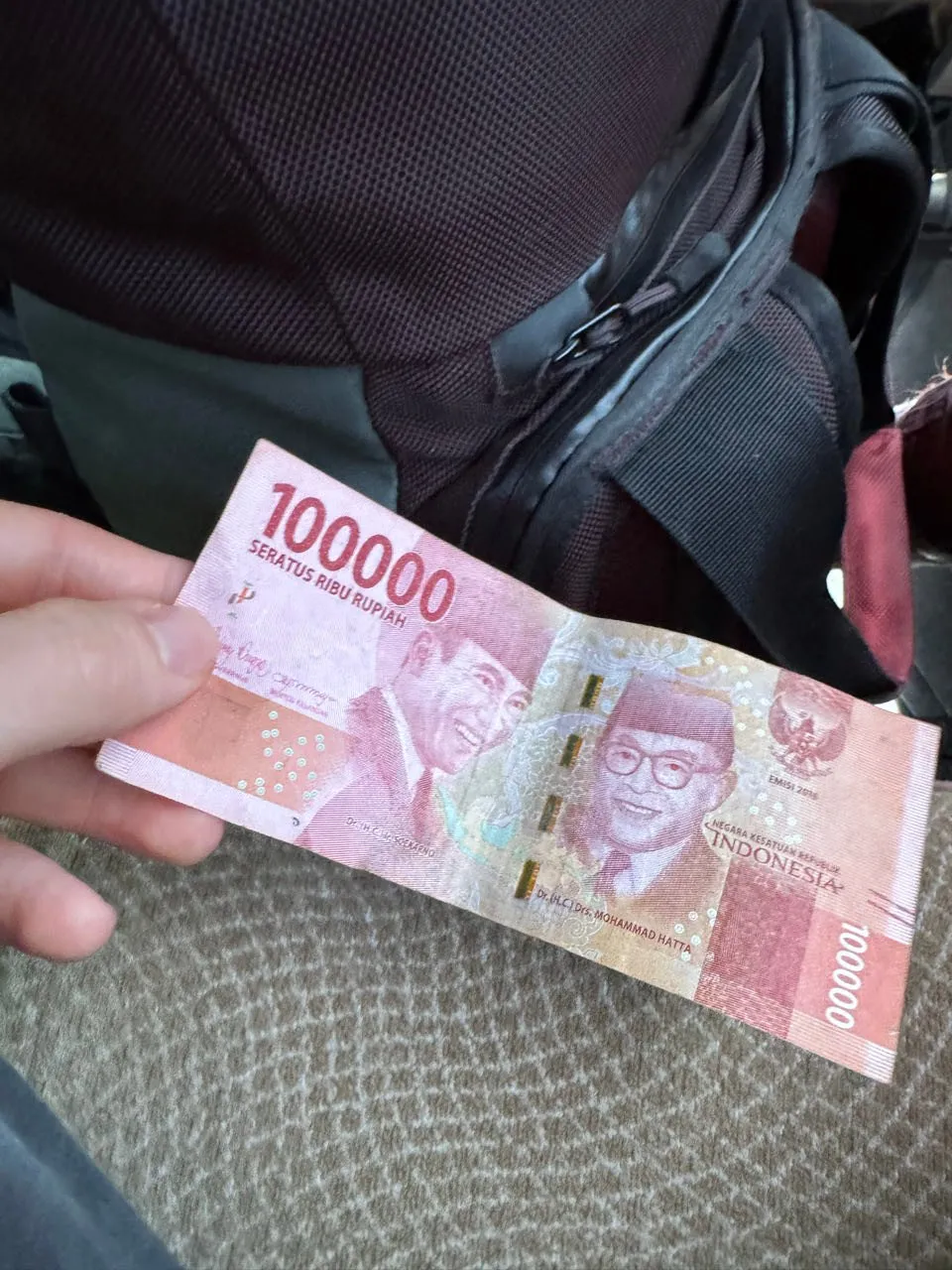 Ein roter Schein über 100'000 Rupiah in der Hand, im Hintergrund ein schwarzer Rucksack