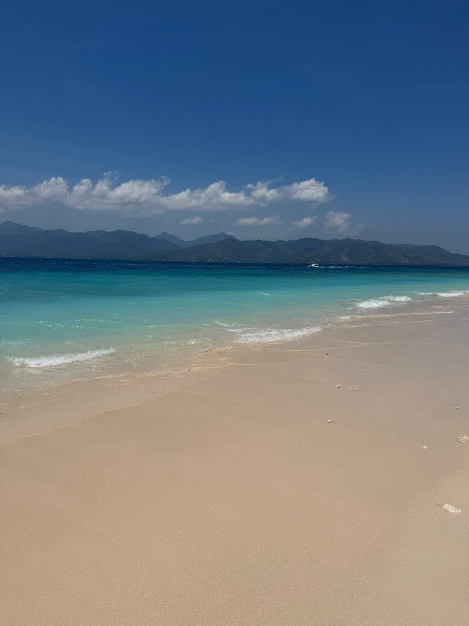 Heller Sandstrand mit sanften Wellen und türkisfarbenem Wasser, am Horizont dunkle Berge unter blauem Himmel