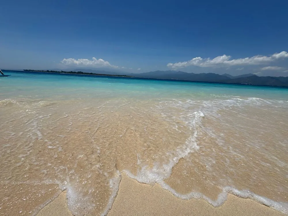 Wellen laufen über hellen Sand ins türkisfarbene Wasser, in der Ferne eine Insel und Berge