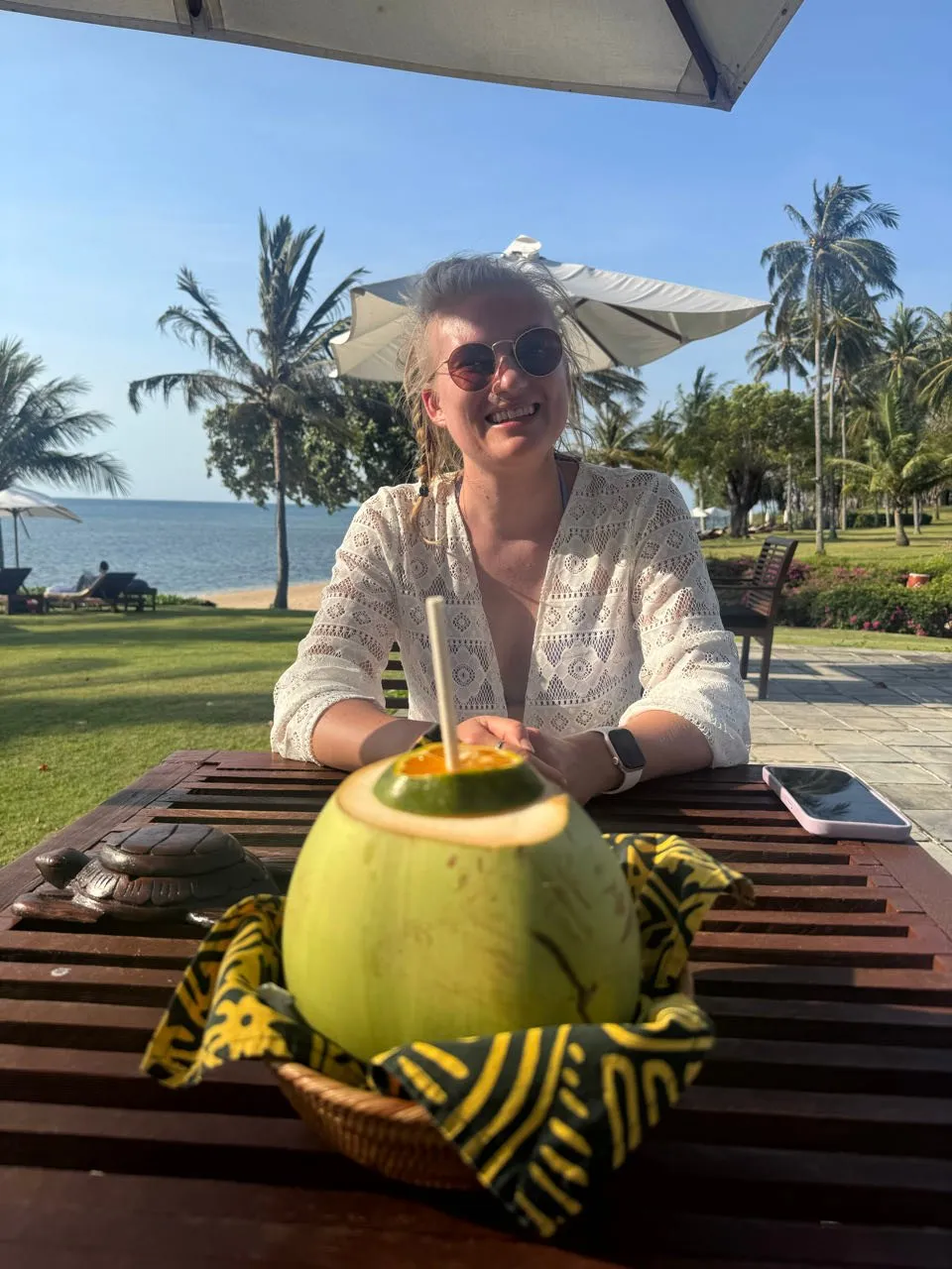 Annina mit Sonnenbrille lächelt an einem Holztisch, vor ihr eine grüne Kokosnuss mit Strohhalm, dahinter Rasen, Palmen und Meer