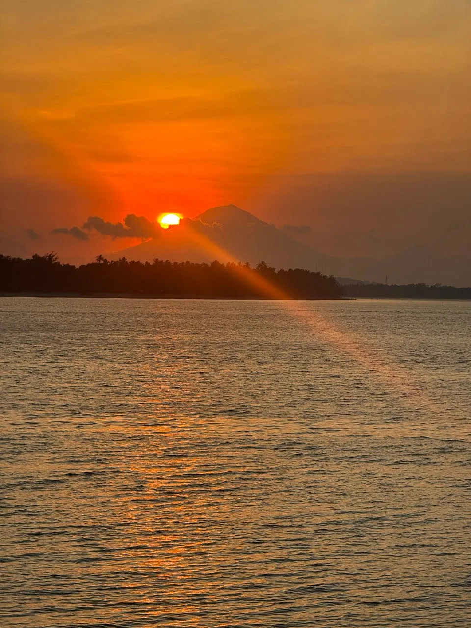 Die Sonne steht als orange Scheibe über einem dunklen Küstenstreifen, dahinter der Kegel eines Vulkans, ein Lichtstrahl fällt über das Wasser