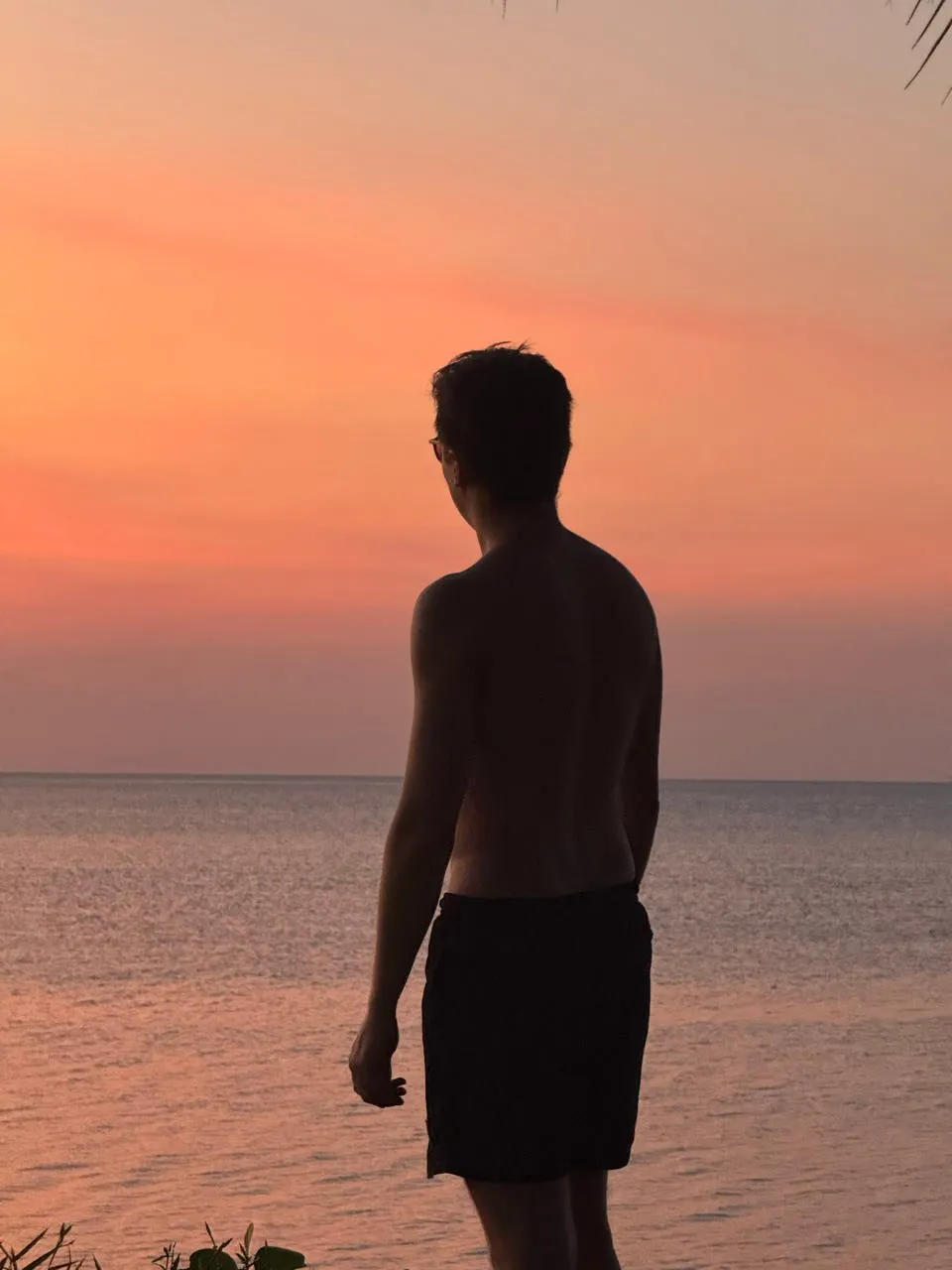 Ein Mann in dunkler Badehose steht als Silhouette am Wasser vor rosa und orangem Himmel